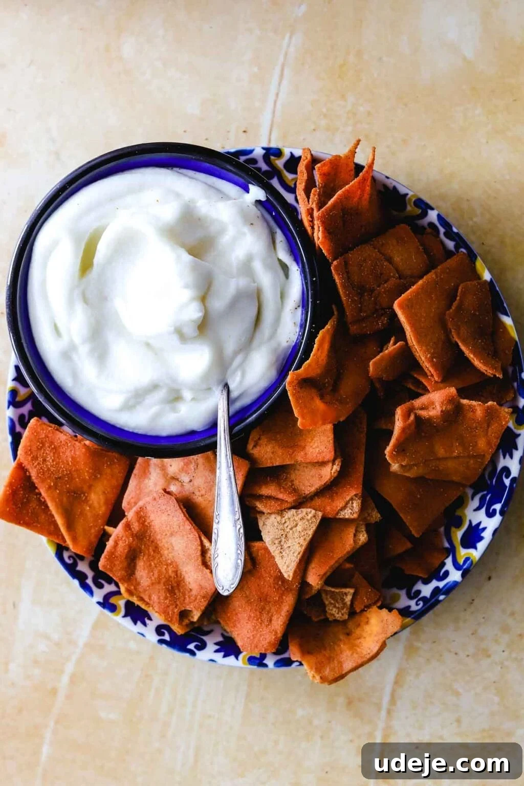 Whip Up Fluffy Toum in 5 Minutes No Food Processor 13 A stylish patterned plate holds golden, crispy pita chips alongside a beautiful bowl of creamy white Toum dip with a spoon, all arranged on a light-colored surface, inviting enjoyment.