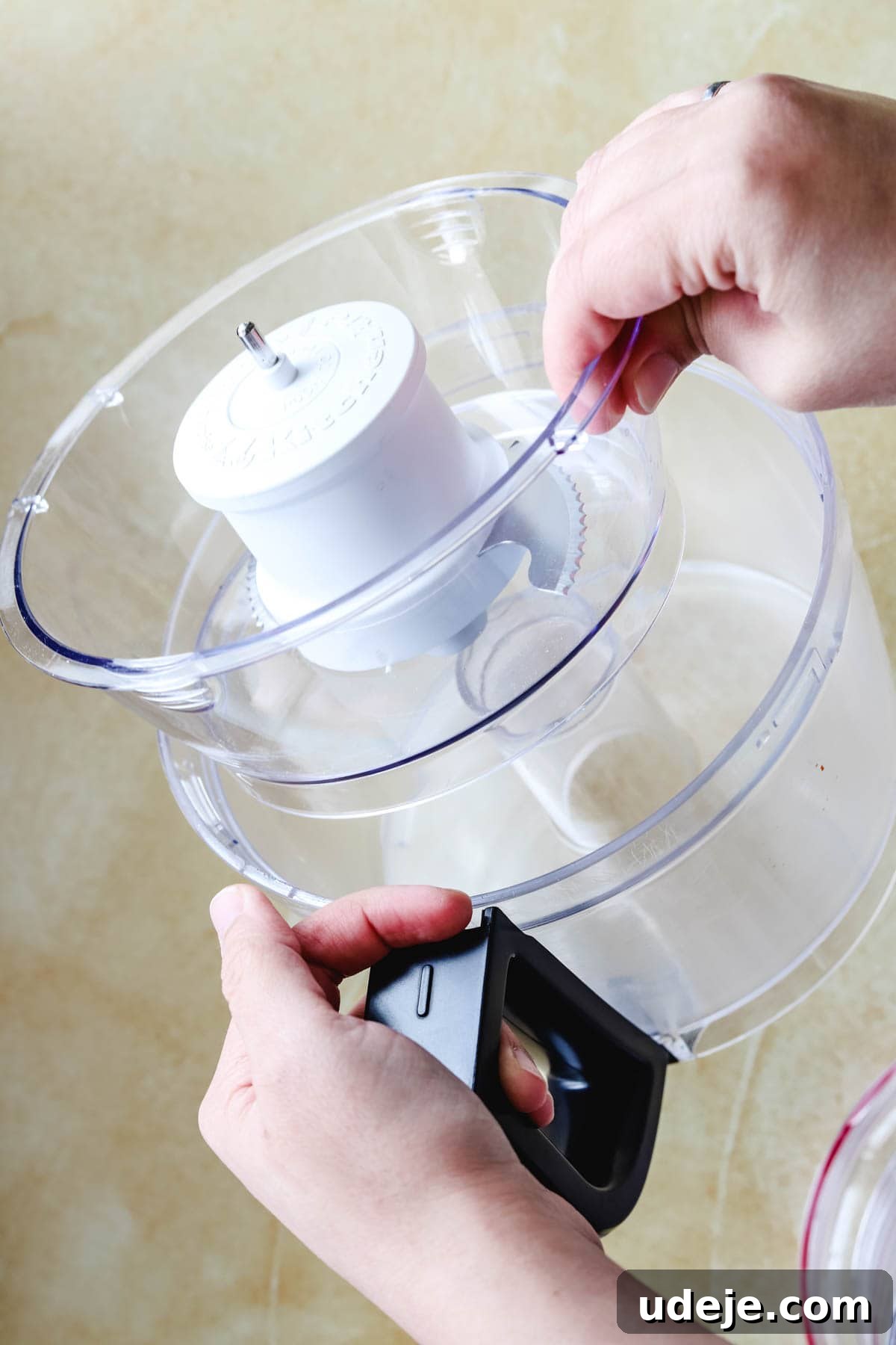 Whip Up Fluffy Toum in 5 Minutes No Food Processor 14 A person’s hands are shown in the process of assembling a clear food processor bowl with a white blade attachment onto its base, in preparation for making fresh Toum, set against a light background.