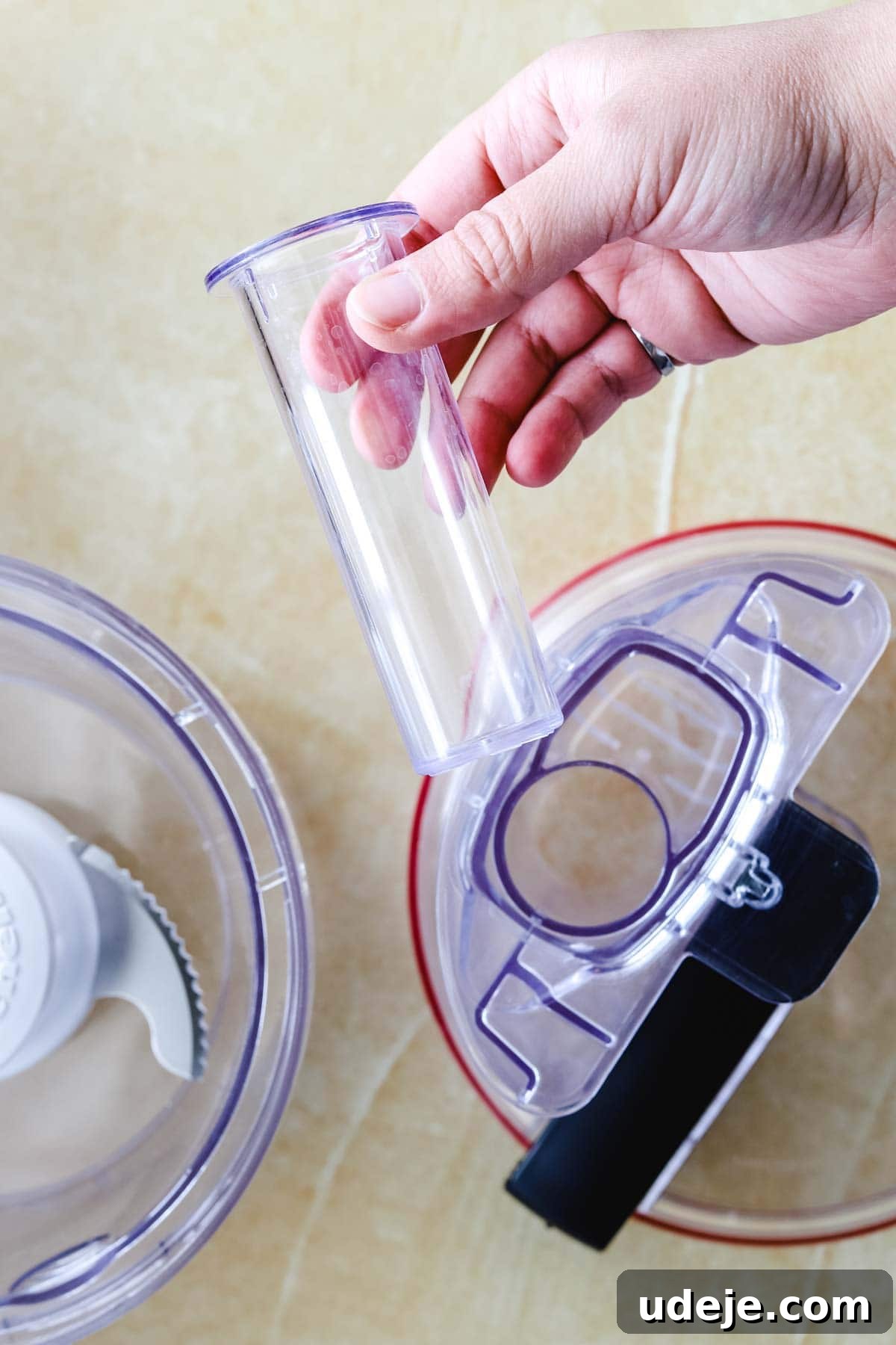 Whip Up Fluffy Toum in 5 Minutes No Food Processor 15 A hand holds a transparent plastic tube over the lid of a food processor, demonstrating an oil streaming attachment, essential for gradually adding oil to create fluffy Toum, on a light-colored surface.