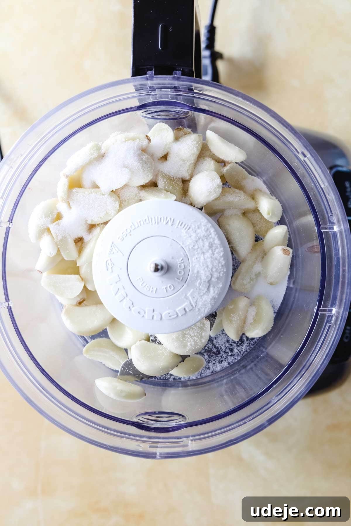Whip Up Fluffy Toum in 5 Minutes No Food Processor 16 An overhead view of a food processor bowl, containing freshly peeled garlic cloves and a dash of salt, resting on a light-colored surface, poised for the creation of classic Toum.