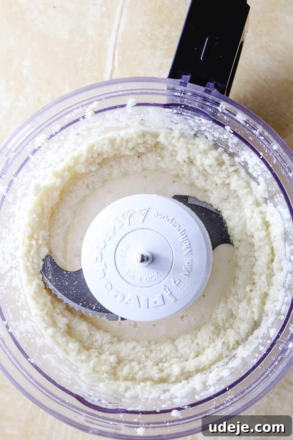 Whip Up Fluffy Toum in 5 Minutes No Food Processor 17 A food processor bowl, viewed from above on a light-colored surface, holds creamy, finely blended Toum, indicating the progress of the garlic pureeing stage.