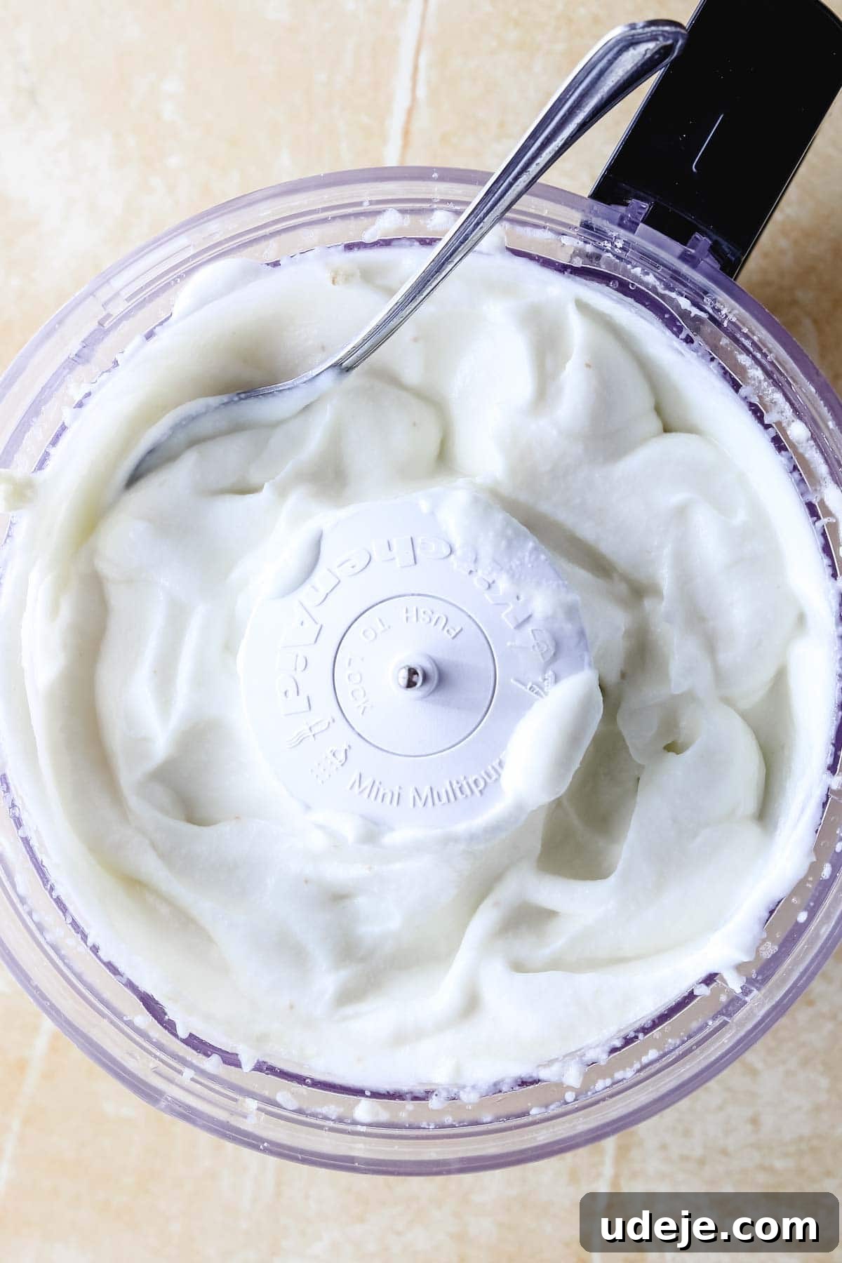 Whip Up Fluffy Toum in 5 Minutes No Food Processor 19 A food processor bowl containing smoothly whipped white Toum, with a metallic spoon resting inside and the processor blade visible in the center, demonstrating a perfectly emulsified garlic sauce.