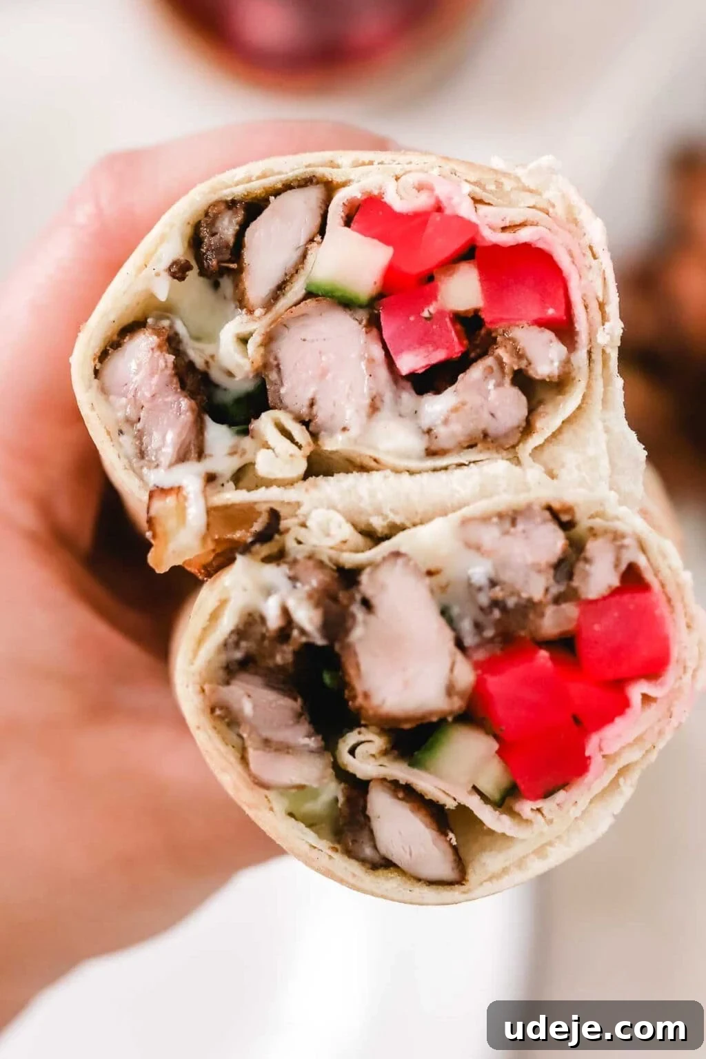 Whip Up Fluffy Toum in 5 Minutes No Food Processor 20 A hand presents a half-cut wrap, showcasing tender chunks of grilled meat, diced red tomatoes, crisp cucumber, and a generous smear of creamy Toum sauce, all nestled within a soft tortilla.