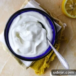 A vibrant blue bowl, filled with creamy white toum sauce and a spoon, rests on stacked napkins, with fresh garlic cloves and half a lemon arranged nearby on a light-colored surface, embodying a fresh and inviting culinary scene.