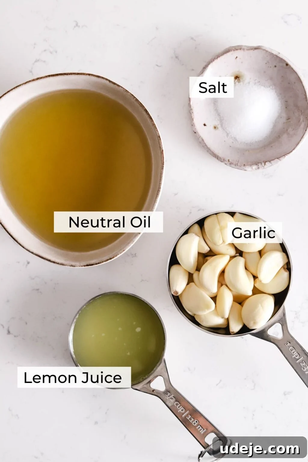 Whip Up Fluffy Toum in 5 Minutes No Food Processor 4 All the essential ingredients for homemade Toum are neatly arranged on a white surface: a bowl of neutral oil, a small dish of salt, a measuring cup brimming with peeled garlic cloves, and a measuring cup of fresh lemon juice. Each element is clearly visible and ready for use.