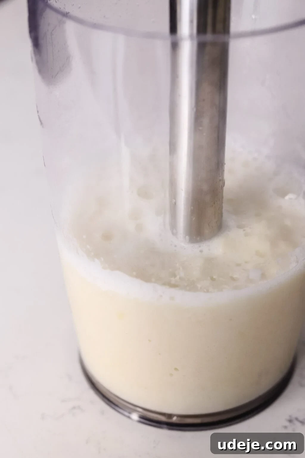 Whip Up Fluffy Toum in 5 Minutes No Food Processor 7 A close-up view of an immersion blender in action, actively mixing a creamy white garlic sauce, Toum, within a clear container, against a light-colored backdrop, highlighting the emulsification process.
