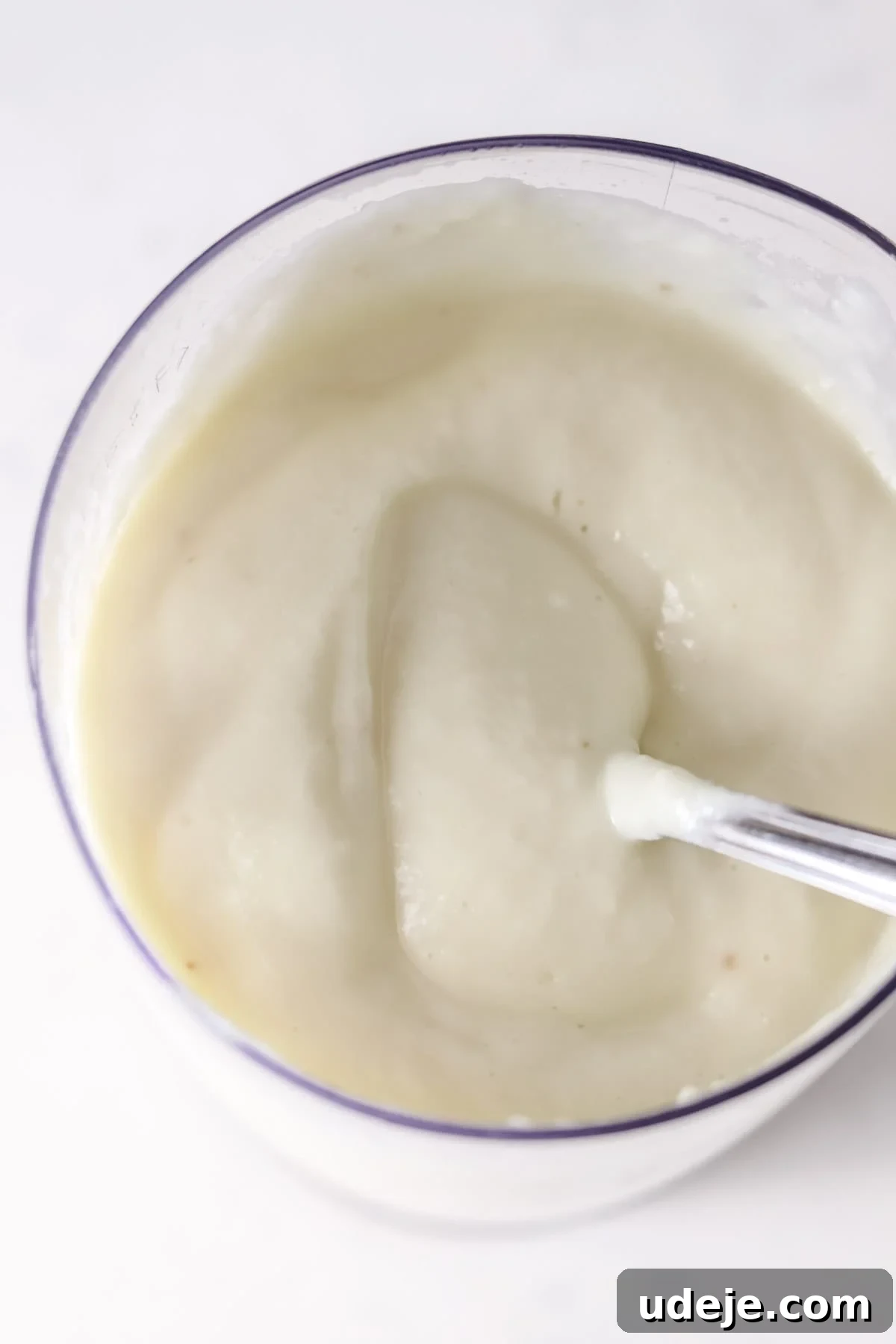 Whip Up Fluffy Toum in 5 Minutes No Food Processor 10 A clear bowl showcasing smooth, creamy Toum, with a metallic spoon partially submerged, reflecting the sauce's thick and glossy texture.