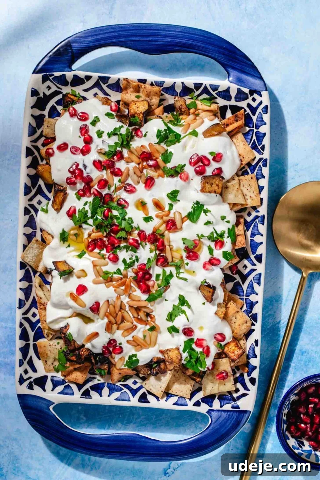 Mediterranean Eggplant Fatteh Delight 2 Eggplant Fatteh served in a patterned blue platter with handles, accompanied by a gold serving spoon and a small blue bowl of pomegranate arils.