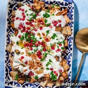 Eggplant fatteh plated on a patterned blue plate with handles, set against a blue backdrop with a gold serving spoon and a small bowl of pomegranate.