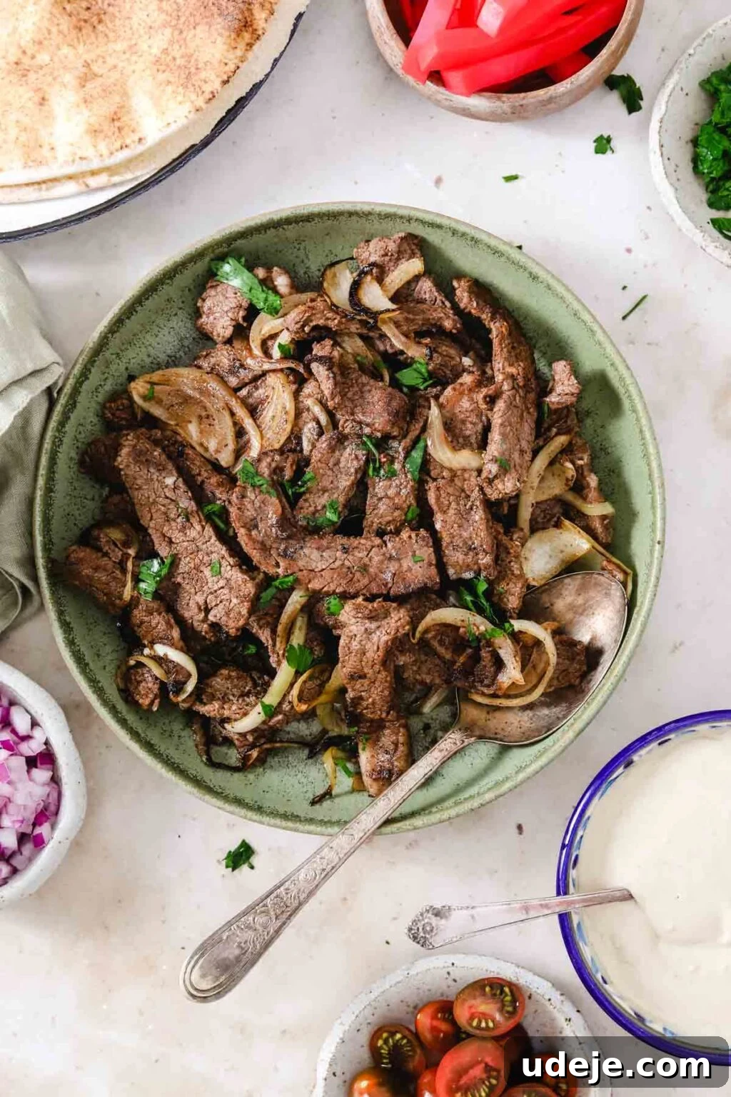 Effortless Skillet Tender Beef Shawarma 2 beef shawarma served in a green bowl with onion slices and chopped parlsey, along with bowls of tomato, tahini sauce, onions, pickles and pita bread