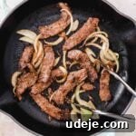 beef shawarma cooked in a cast iron skillet with onion slices and silver tongs