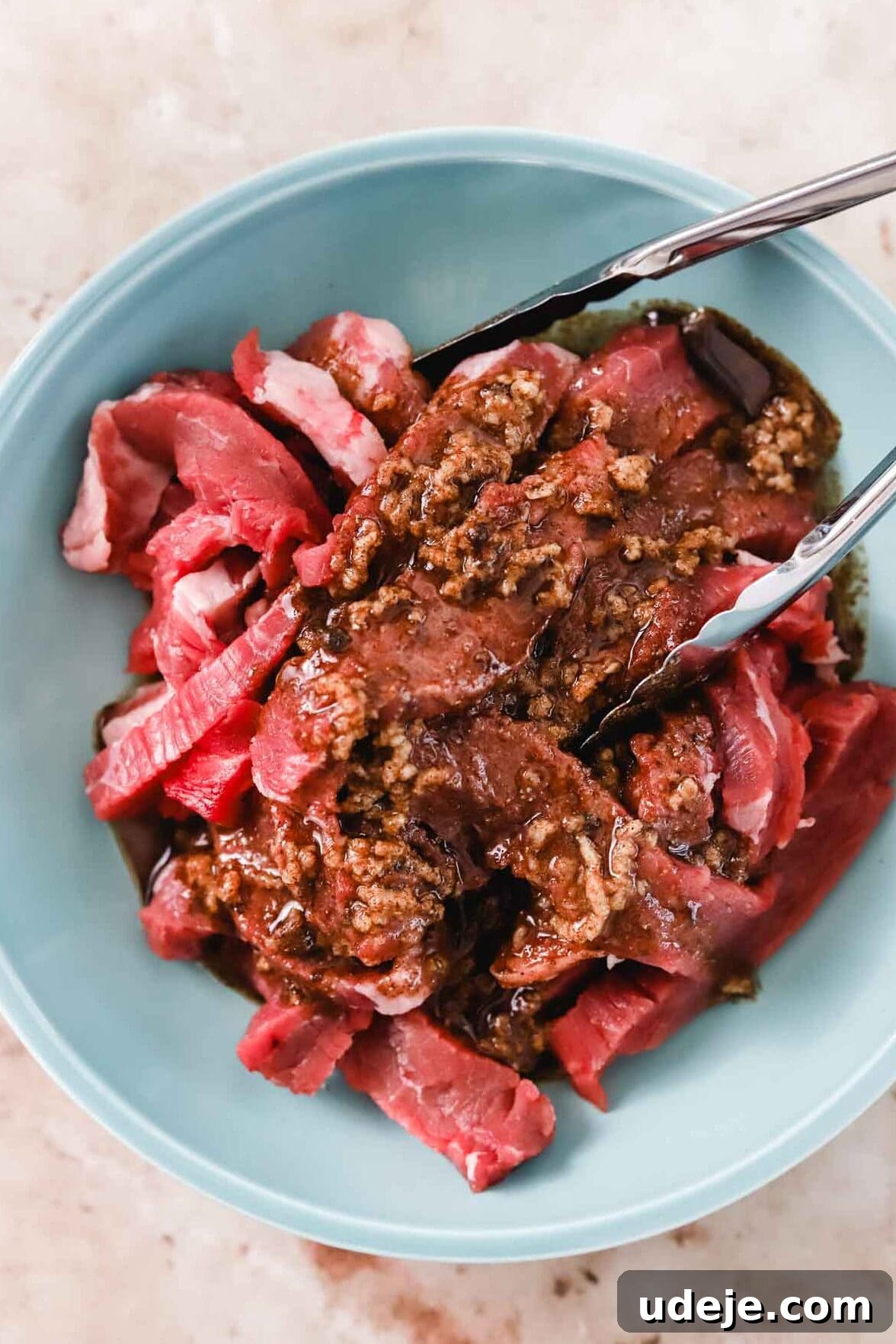 Effortless Skillet Tender Beef Shawarma 6 beef shawarma marinating in a blue bowl with a marinade mixture on top and silver tongs