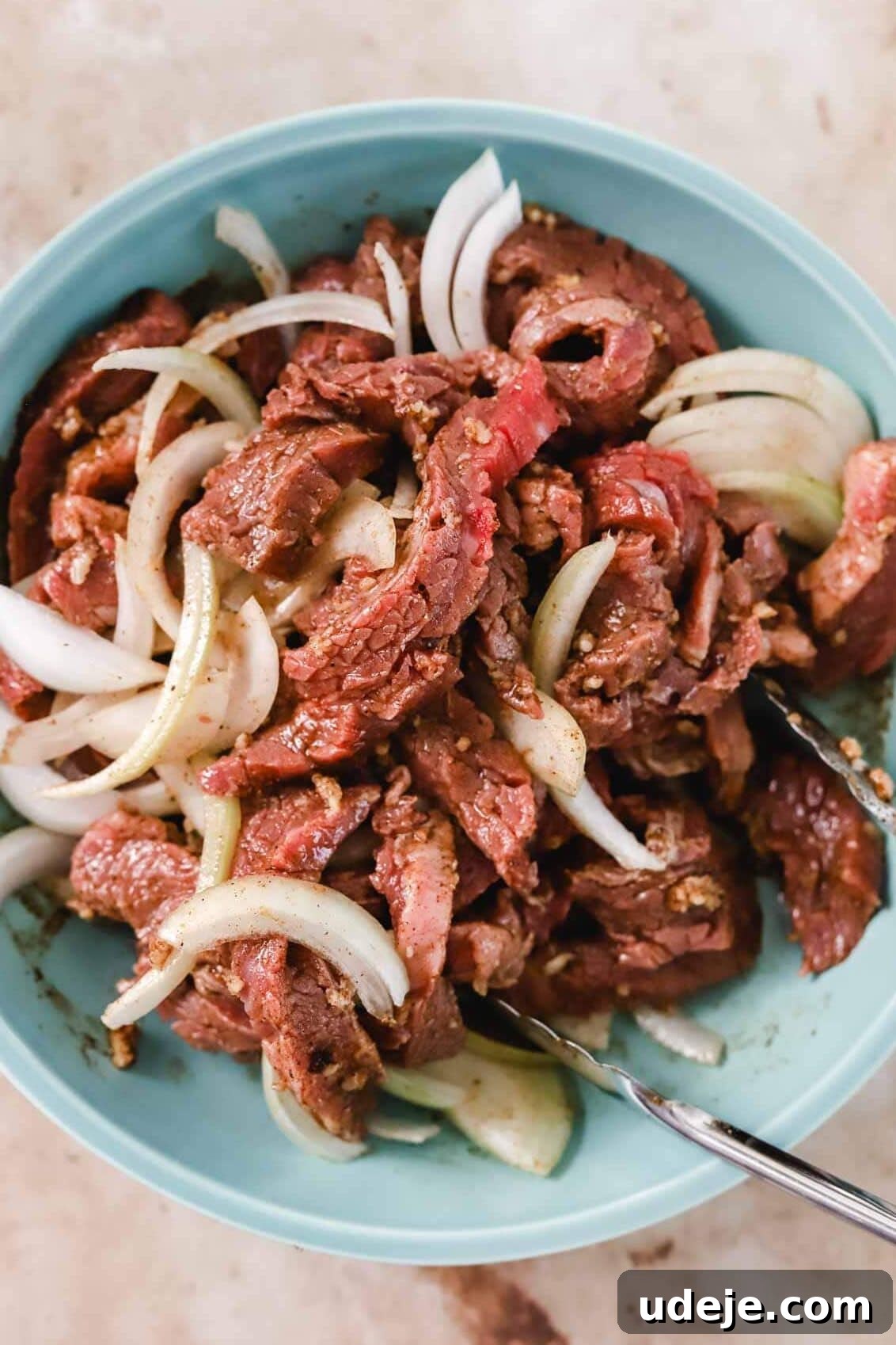 Effortless Skillet Tender Beef Shawarma 7 beef shawarma marinating with onions slices in a blue bowl with silver tongs