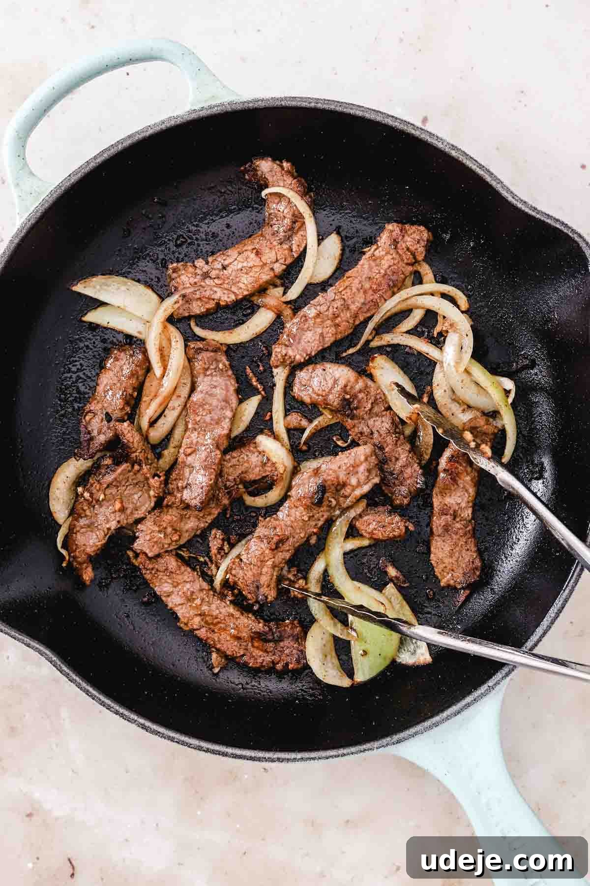 Effortless Skillet Tender Beef Shawarma 8 beef shawarma cooked in a cast iron skillet with onion slices and silver tongs