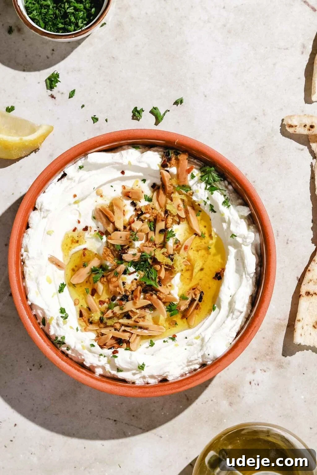 Orange-brown bowl filled with labneh topped with slivered almonds, parsley, with pita at the side, olive oil bottle, parsley and lemon at the side