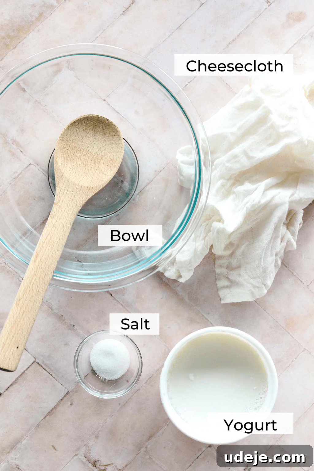 Ingredient shot for Labneh with a medium sized glass bowl with a wooden spoon inside, a small glass bowl with salt, a yogurt container and cheesecloth on a light pink background