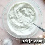 medium sized glass bowl with yogurt and a spoon, small glass bowl with salt, wooden spoon and cheesecloth in the corners on a pink background