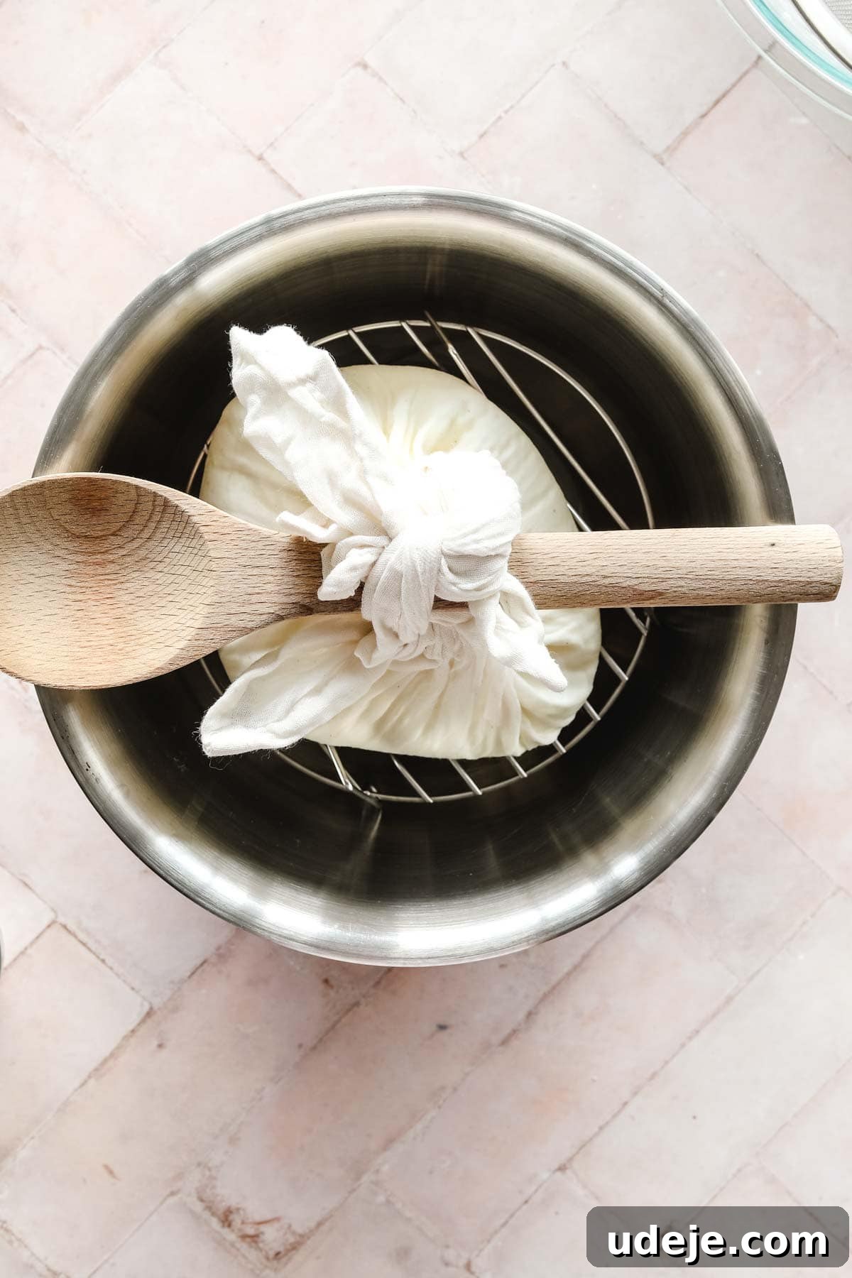 steel bowl with cheesecloth tied to wooden spoon on a trivet, on a pink background