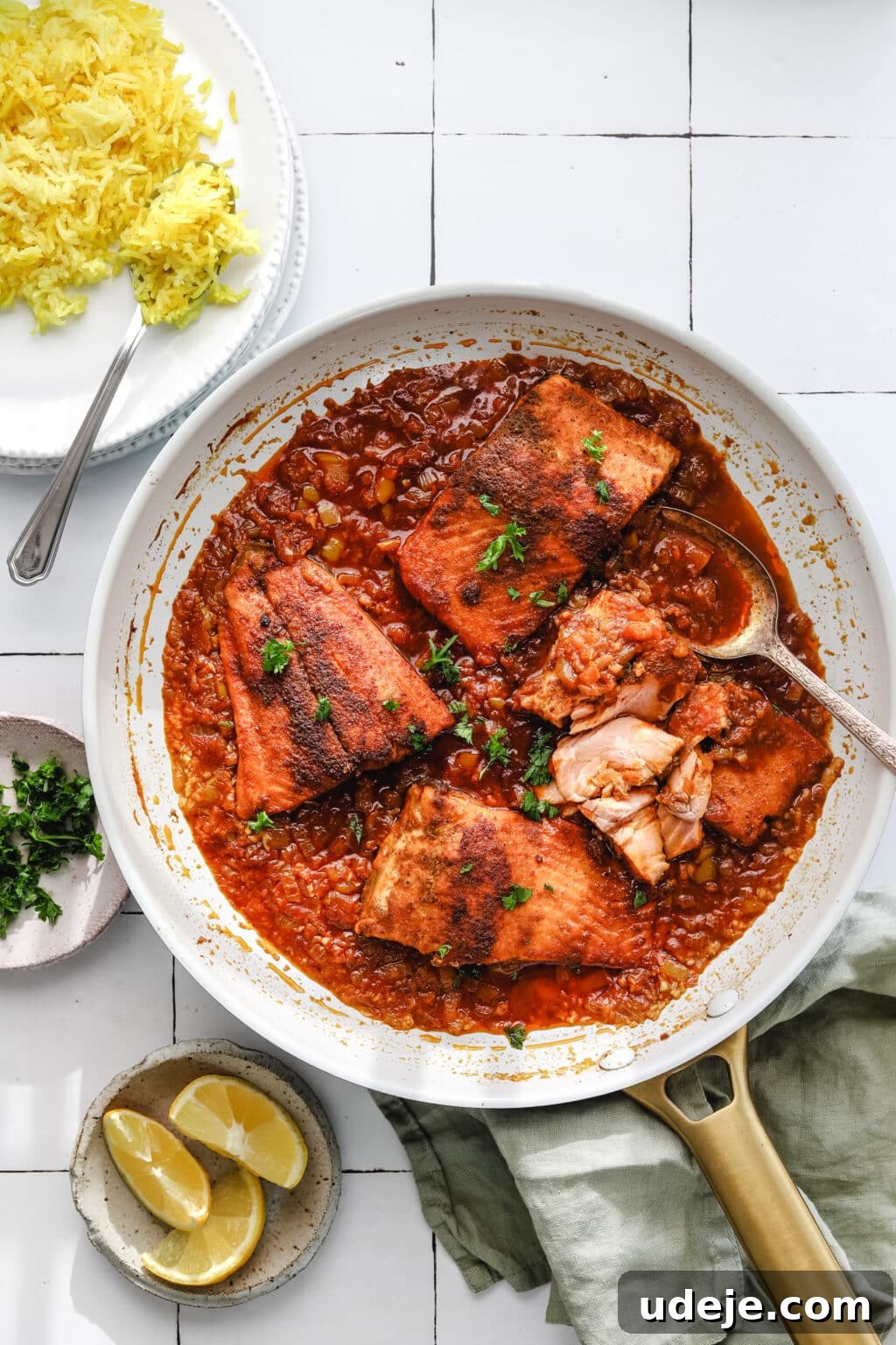 A light-colored skillet filled with perfectly cooked fish pieces simmering in a rich, vibrant tomato sauce, artfully arranged on a white tiled background. In the corner, a platter of golden yellow rice awaits, accompanied by a small bowl of fresh parsley, bright lemon slices, and a neatly folded green napkin, inviting you to a delightful Mediterranean feast.