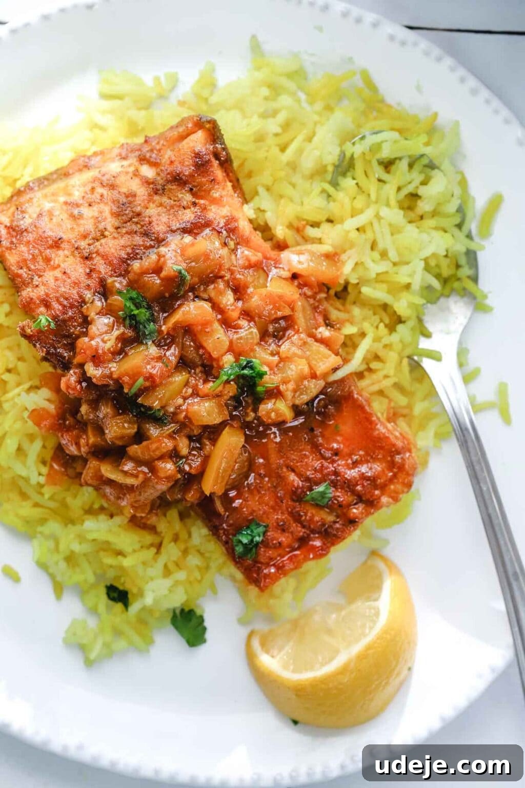 A perfect piece of Mediterranean fish, bathed in vibrant tomato sauce, rests atop a mound of fragrant yellow rice on a pristine white plate. A fork is poised nearby, ready for indulgence, alongside a bright lemon wedge, offering a burst of freshness.