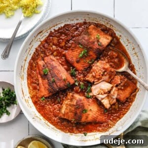 A light-colored skillet brimming with pieces of fish in a rich tomato sauce, positioned beside a platter of golden yellow rice. A small bowl of fresh parsley, bright lemon slices, and a green napkin are elegantly arranged on a white tiled background, creating a mouth-watering scene.
