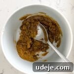 A small white bowl containing a vibrant, golden spice mix, with a spoon resting inside, ready for marination on a light-colored background.