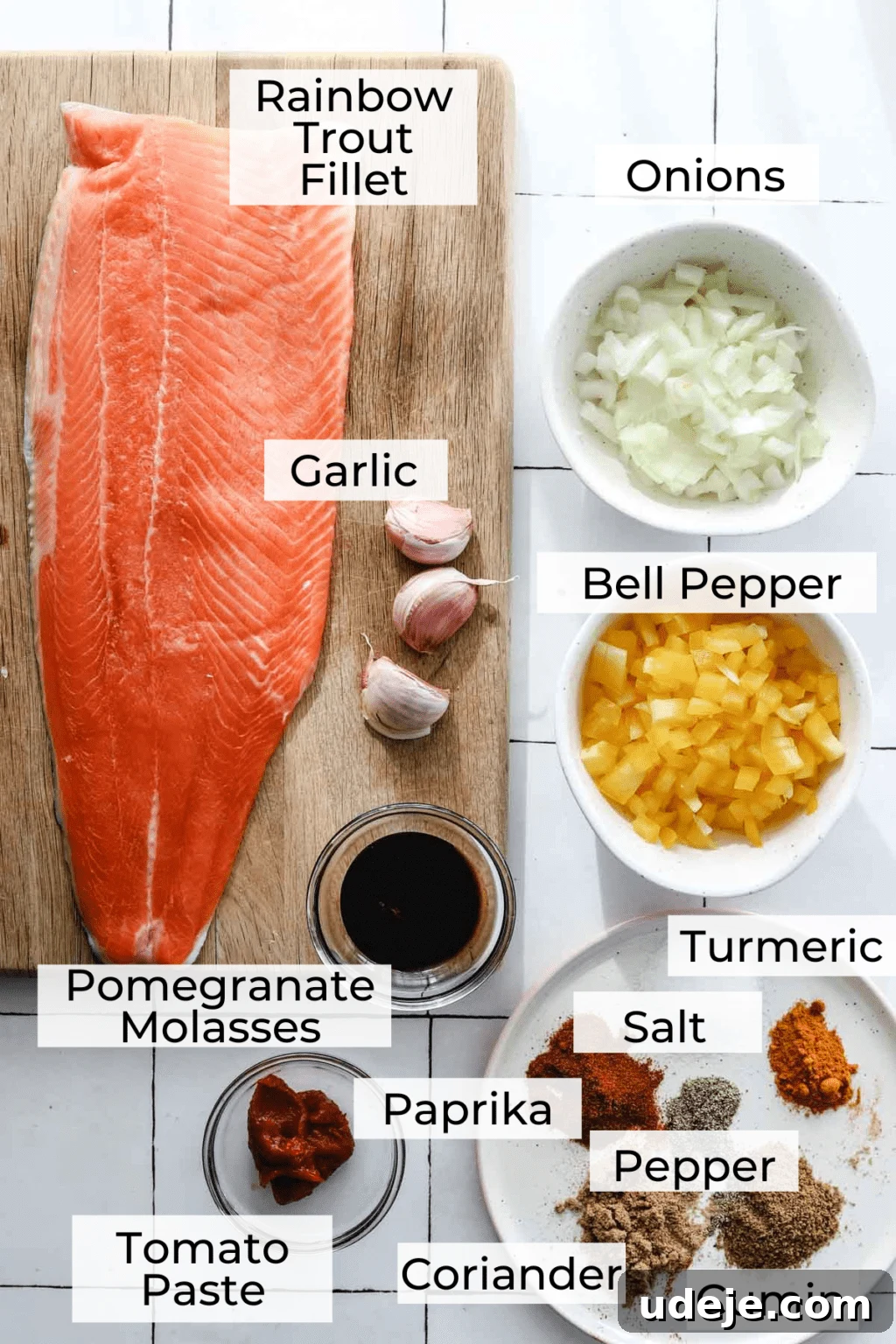 A vibrant display of fresh ingredients for Mediterranean fish: a glistening rainbow trout fillet, plump garlic cloves, and a rich bottle of pomegranate molasses arranged on a rustic wooden cutting board. Alongside, crisp onions and bright yellow bell peppers are nestled in white bowls, an array of colorful spices are neatly presented on a small plate, and concentrated tomato paste sits ready in a tiny bowl, all set on a pristine white tile background.