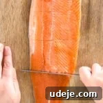 A skilled hand carefully cuts a rainbow trout fillet horizontally into equal pieces on a rustic wooden cutting board, with another hand steadying the fish, ensuring precise and even portions.