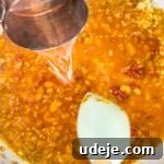 A measuring cup pours hot water into a light-colored skillet, where a rich tomato sauce is forming, being stirred by a white spatula to create a perfect consistency.