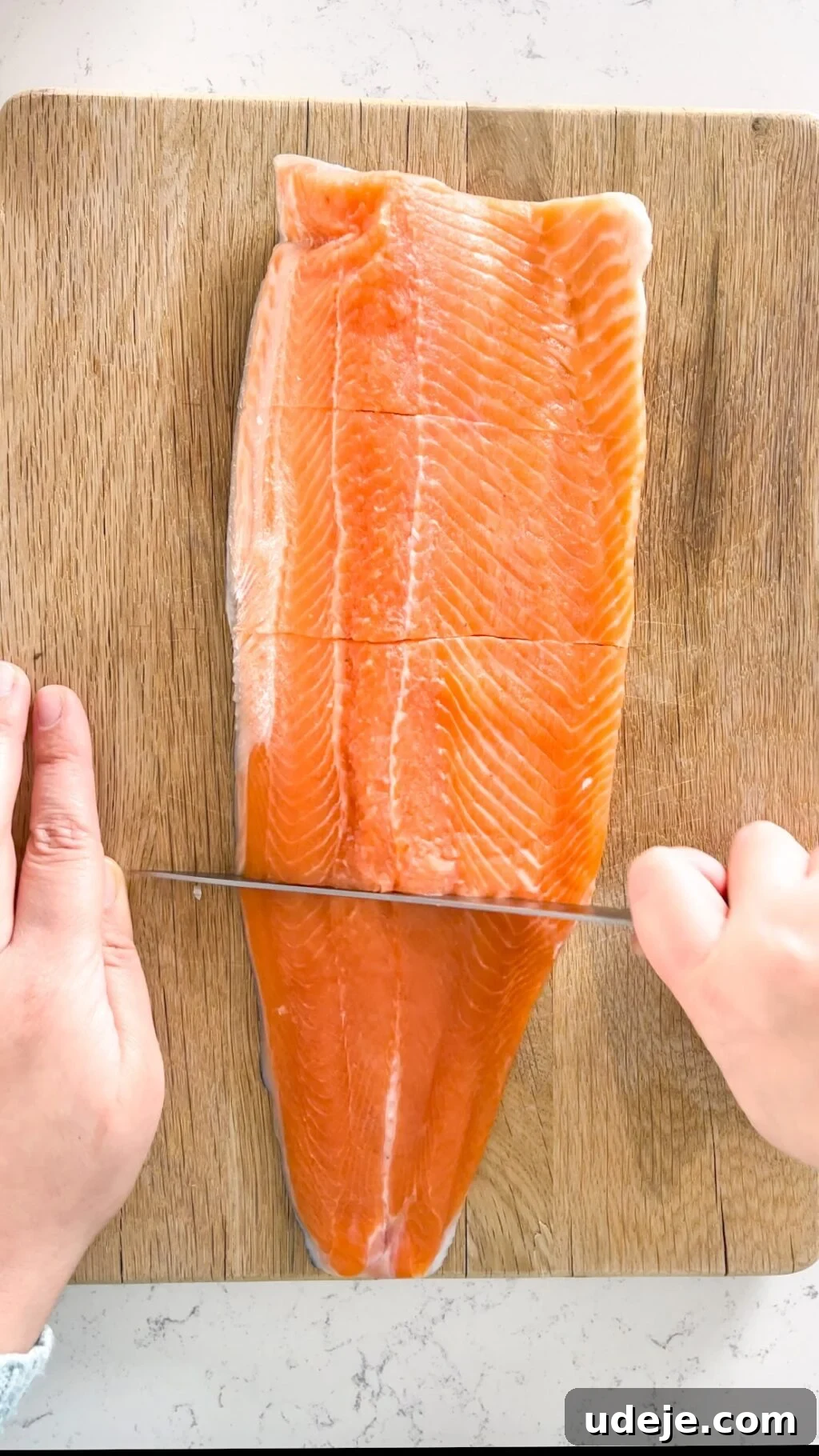 A skilled hand carefully cuts a rainbow trout fillet horizontally into equal pieces on a rustic wooden cutting board, with another hand steadying the fish, ensuring precise and even portions.