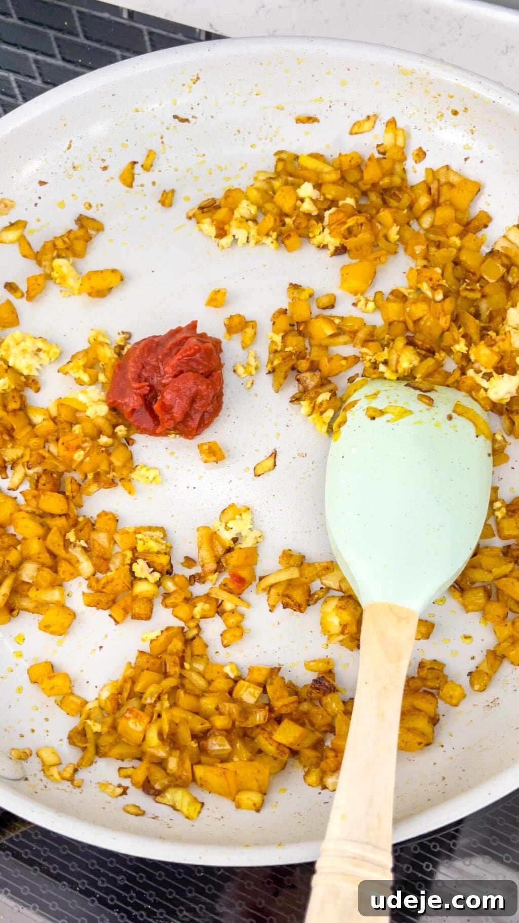 A light-colored skillet filled with beautifully cooked onions and vibrant yellow peppers, mingling with rich tomato paste, as a white spatula gently stirs the aromatic mixture.