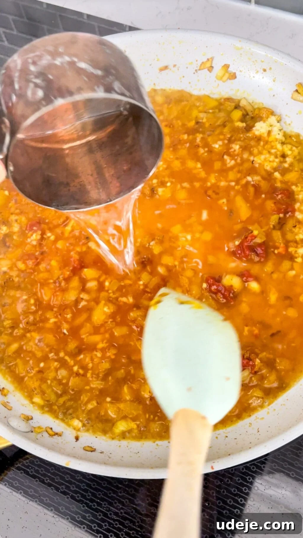 A measuring cup pours hot water into a light-colored skillet, where a rich tomato sauce is forming, being stirred by a white spatula to create a perfect consistency.
