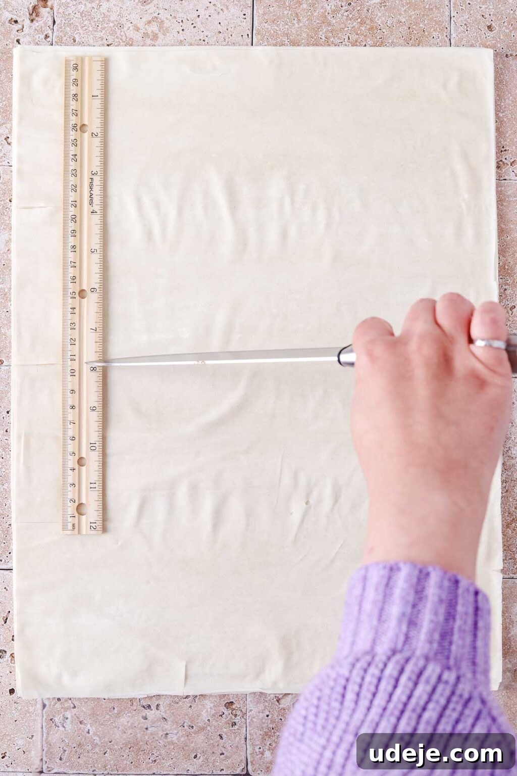 A phyllo sheet on a light stone tiled background, being cut horizontally with a knife using a ruler as a guide.