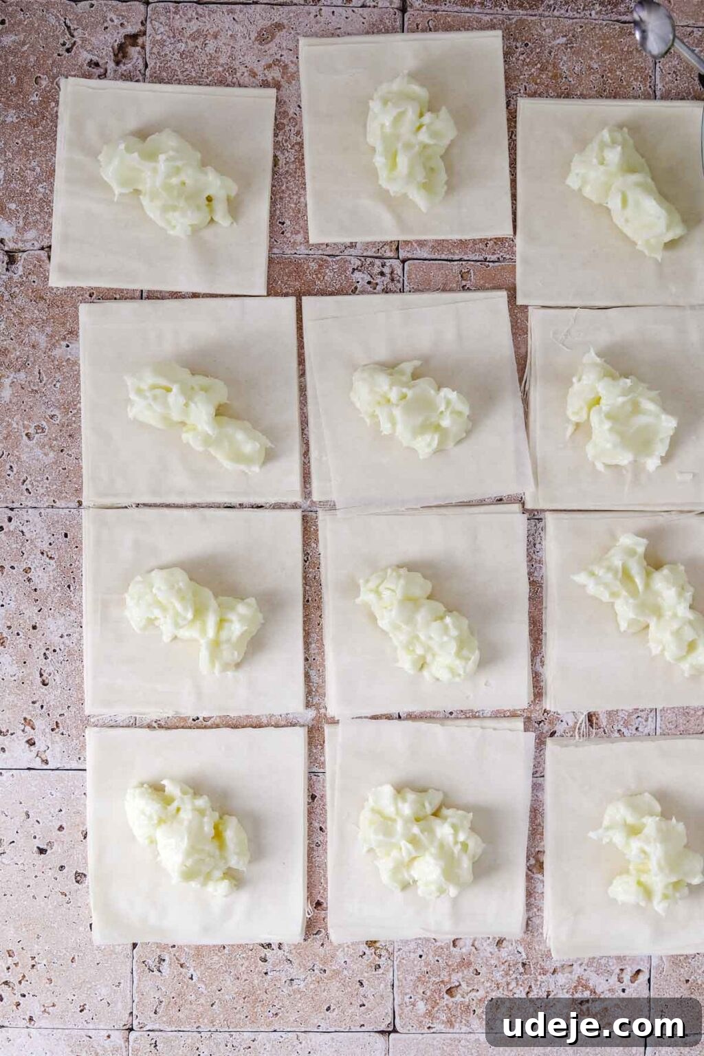 12 cut pieces of phyllo dough with a dollop of ashta cream placed in the middle of each, arranged on a light stone tiled background.