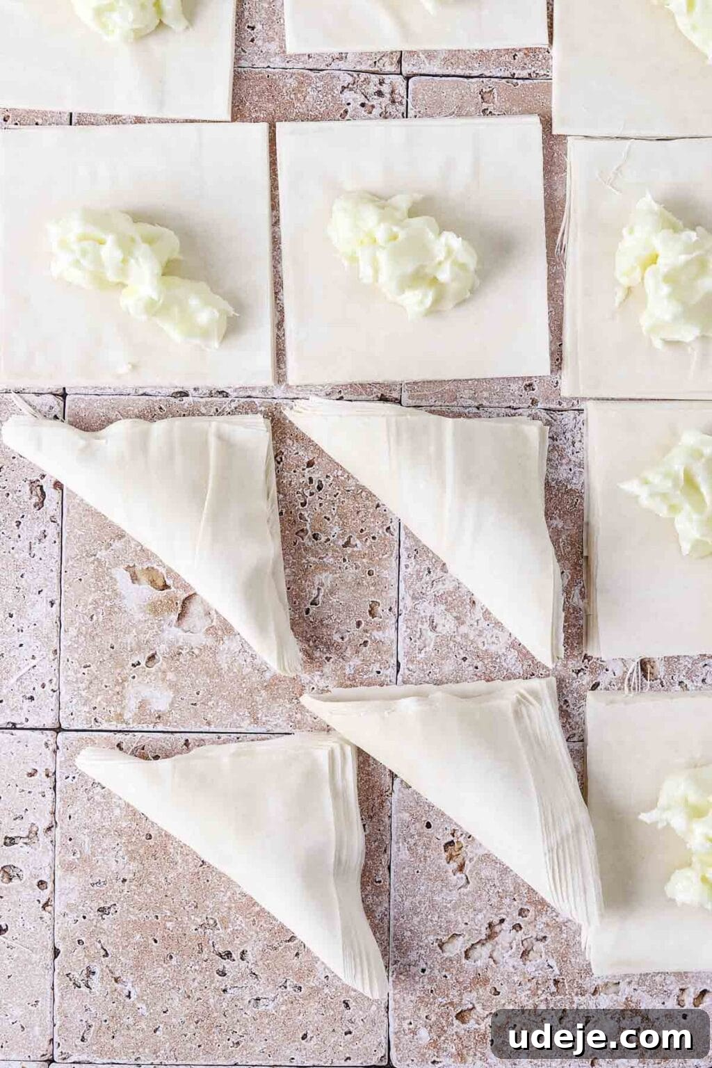Pieces of square-cut phyllo with ashta in the middle, with four of them already folded into triangles, all on a light stone tiled background.