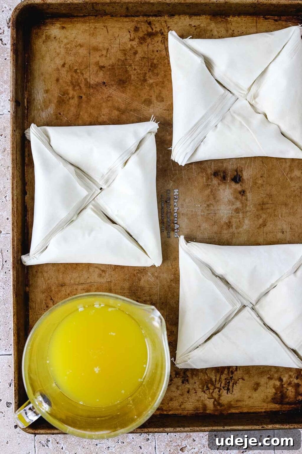 Three squares, each composed of four triangular Warbat pieces, arranged on a baking sheet with a glass measuring cup of melted butter also on the sheet, all on a light stone tiled background.