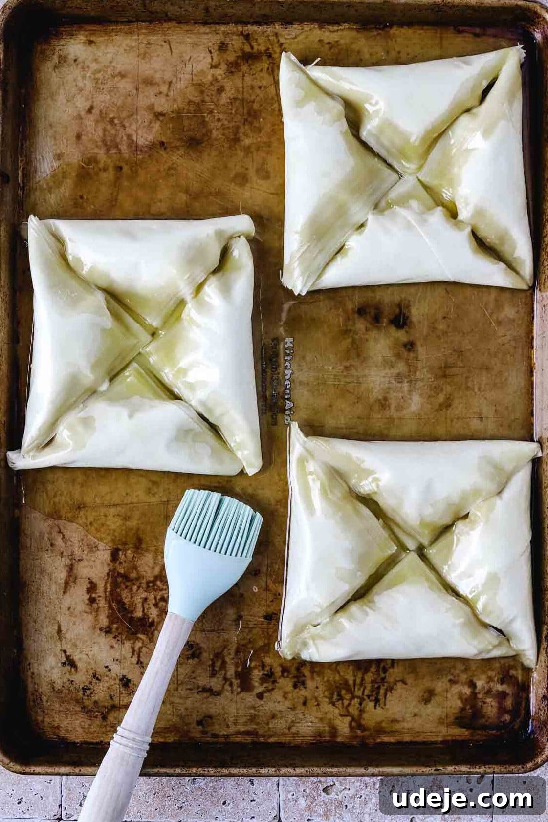 Three squares, each made of four triangular Warbat pieces, on a baking sheet, brushed thoroughly with butter using a silicone brush, on a light stone tiled background.