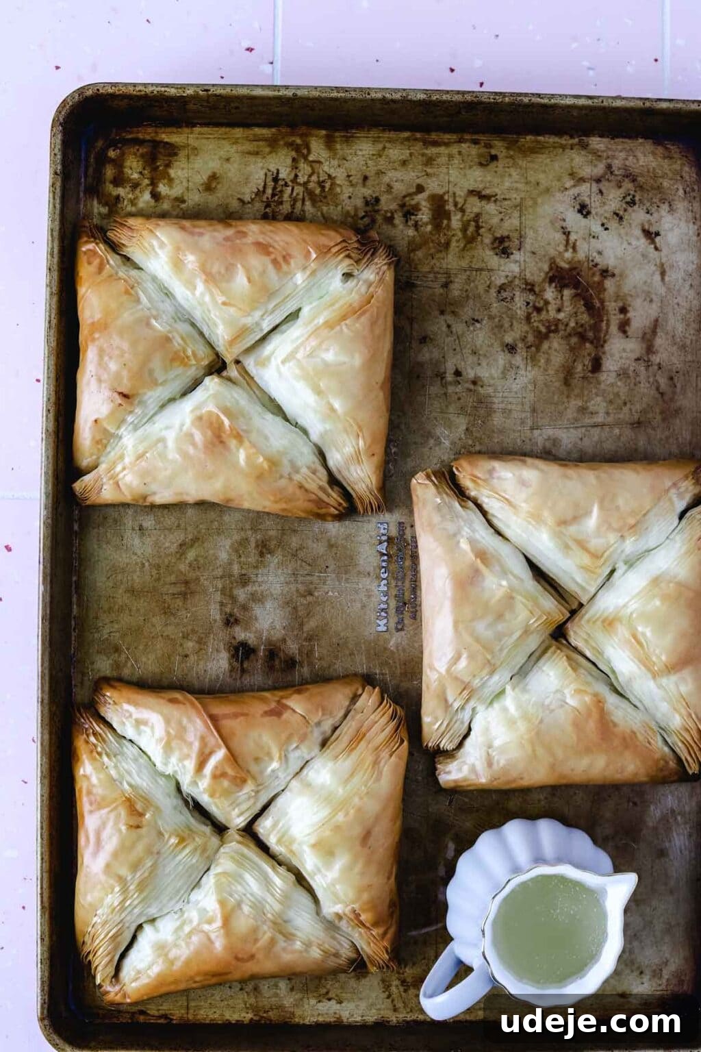 Three squares, each made of four triangular Warbat pieces, on a baking sheet after being baked, with a small pitcher of sugar syrup nearby.
