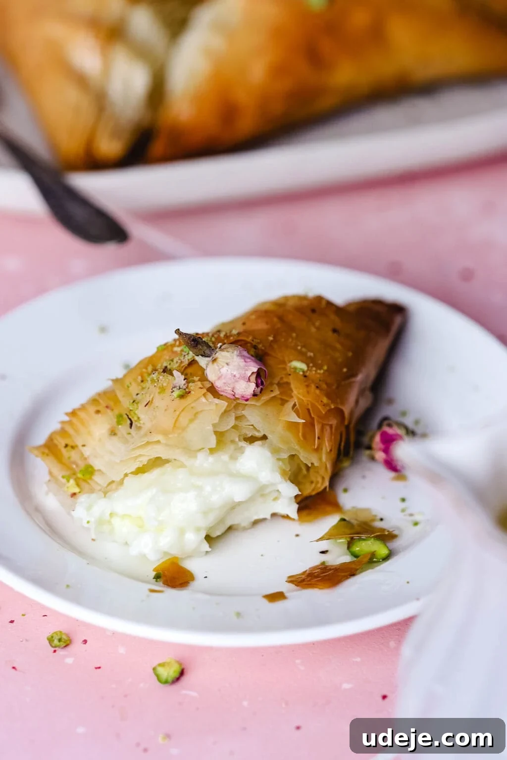 A Warbat pastry on a white plate with a piece torn off, revealing its creamy filling. More Warbat pieces are visible on a serving platter in the background, adorned with crushed pistachios and rose petals, set against a vibrant pink tiled surface.