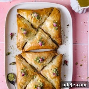 Two squares of Warbat pastries on a rectangular white plate, garnished with crushed pistachios and delicate rose petals. A small spoon and a bowl of pistachios are in the corner, with a teacup, saucers, and sugar syrup pitcher in the background on a bright pink tiled surface.