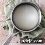 A silver saucepan with water and sugar simmering on a stovetop, resting on a light green tea towel against a stone background.