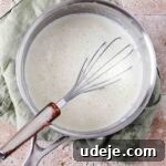 A silver saucepan holding a cream mixture, with a whisk submerged in it, resting on a light green tea towel on a stone tiled background.