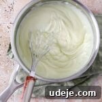 A silver saucepan with thickened ashta and a whisk, resting on a light green tea towel on a stone tiled background.