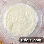 Ashta covered with plastic wrap, pressed onto its surface in a glass bowl, resting on a light stone tiled background.