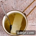 A small gold saucepan with a stick of butter and melted butter, with a spoon sticking out, on a light stone tiled background.