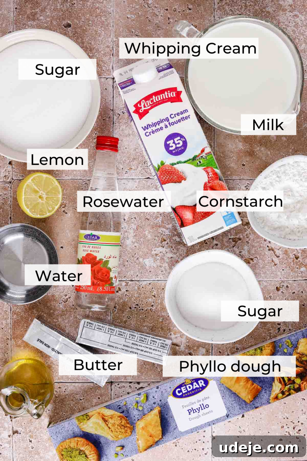 Ingredients for Warbat laid out: a medium white bowl of sugar, a smaller white bowl of sugar, a small white bowl of cornstarch, a measuring cup with milk, a whipping cream container, a bottle of rosewater, water in a silver measuring cup, sticks of butter, and a phyllo dough package, all arranged on a light stone tiled background.