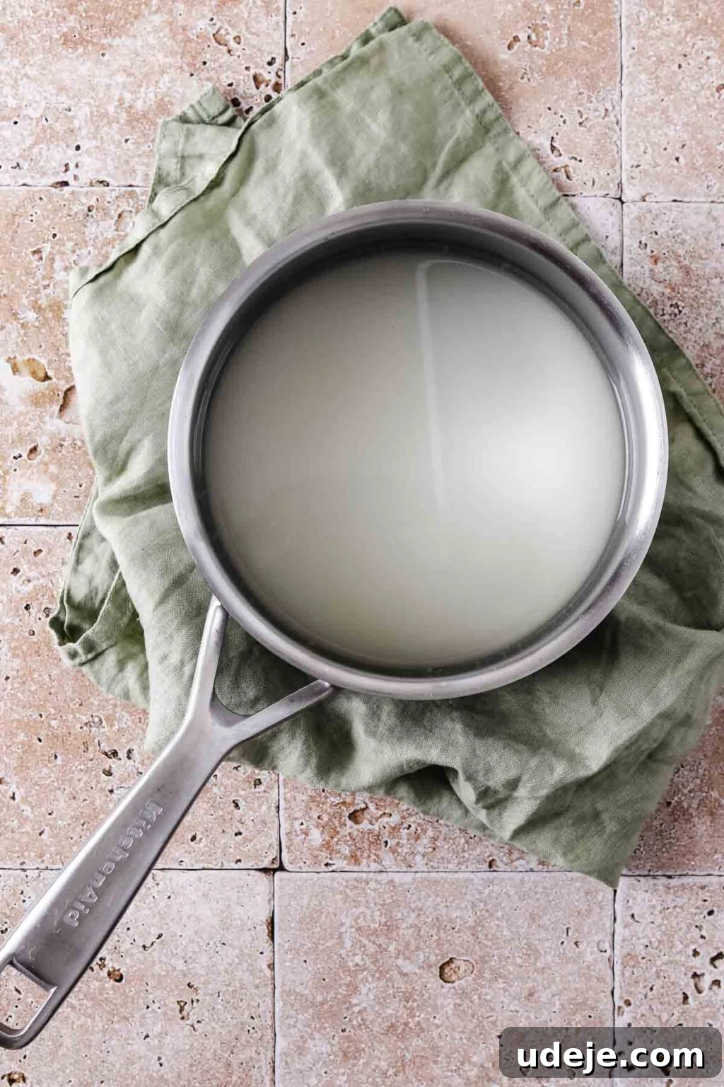 A silver saucepan with water and sugar simmering on a stovetop, resting on a light green tea towel against a stone background.