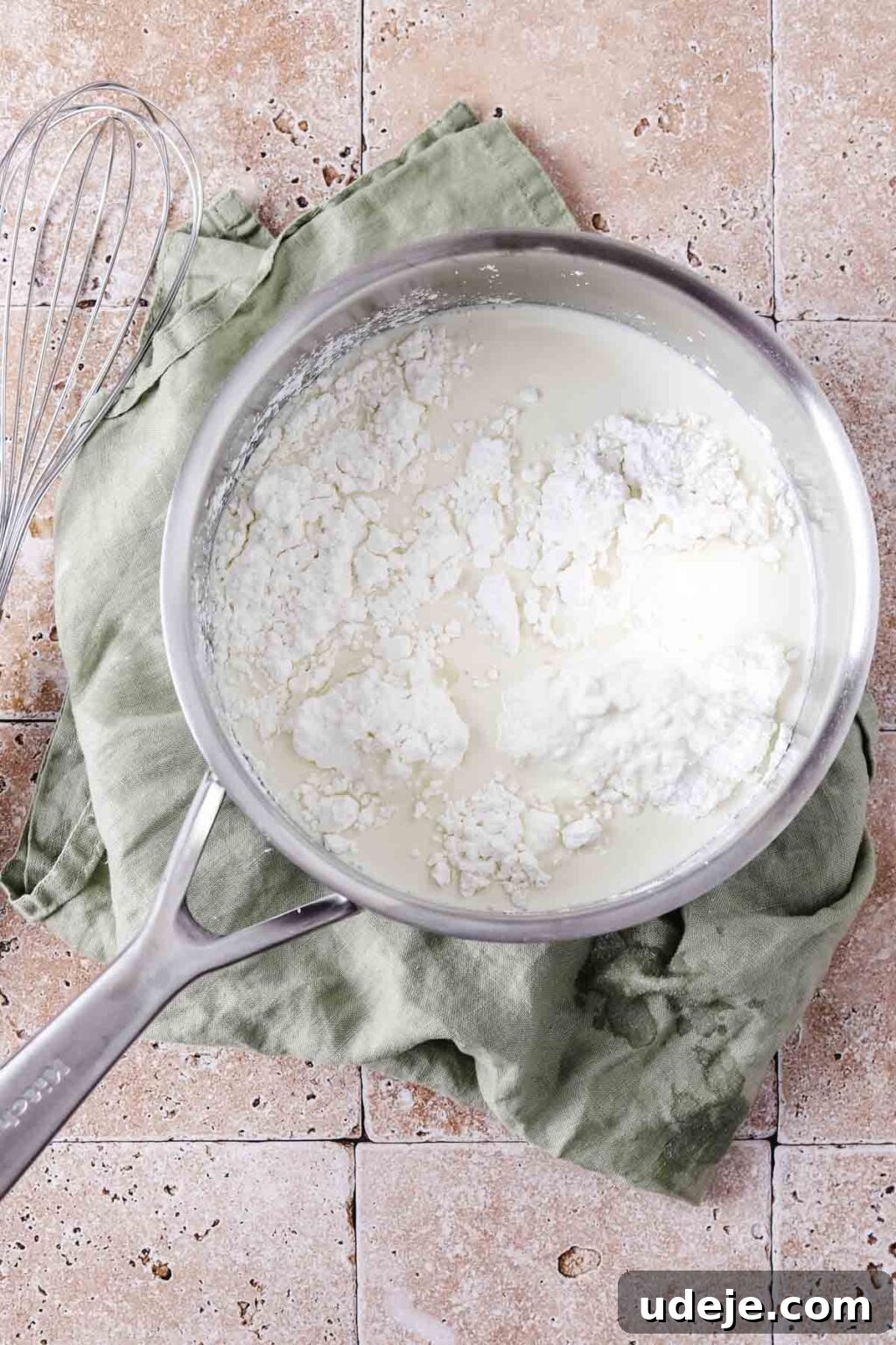A silver saucepan containing cream and cornstarch, with a whisk beside it on a light green tea towel on a stone tiled background.