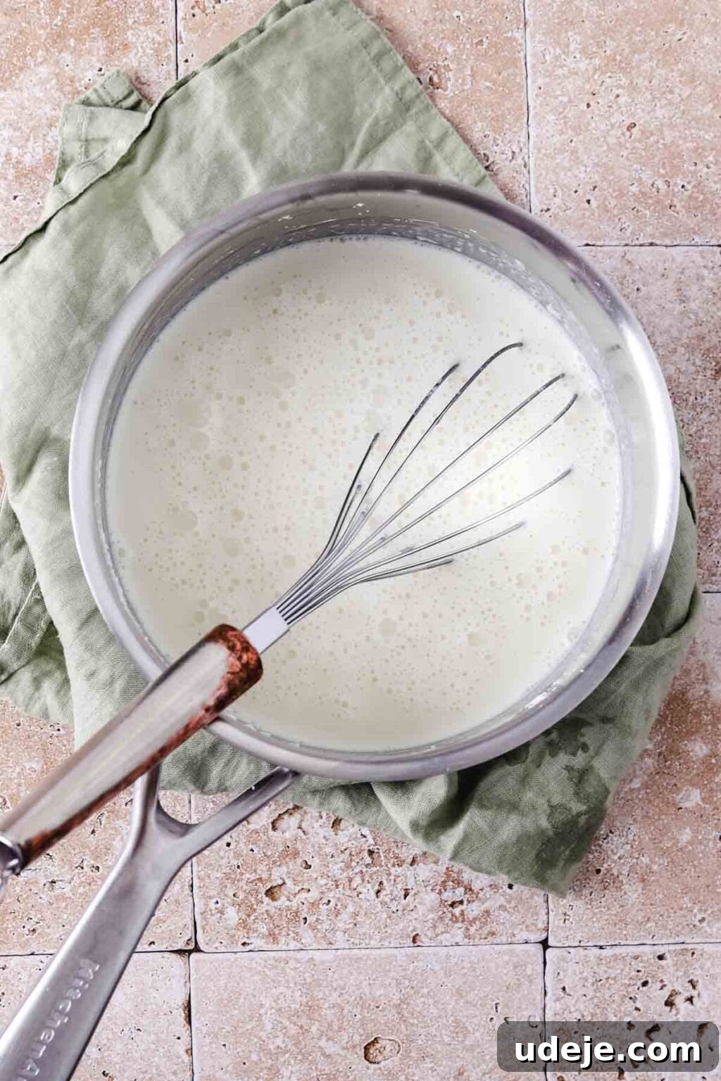 A silver saucepan holding a cream mixture, with a whisk submerged in it, resting on a light green tea towel on a stone tiled background.