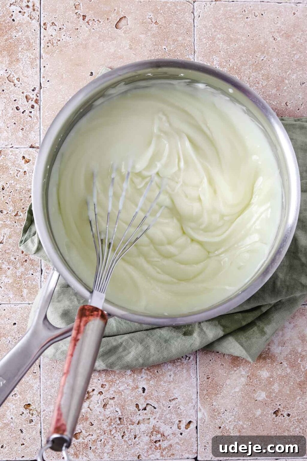 A silver saucepan with thickened ashta and a whisk, resting on a light green tea towel on a stone tiled background.